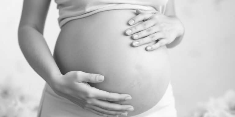 A black-and-white image of a pregnant person gently cradling their belly with both hands. They are dressed in a light-colored top and pants, evoking thoughts of the journey ahead, such as prenatal DNA testing. The background is softly blurred, adding to the serene atmosphere.